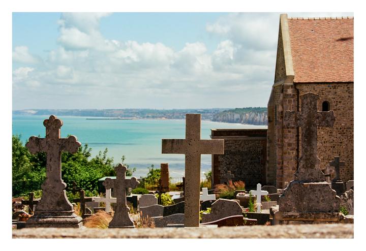 A graveyard with many crosses and tombstones overlooks a body of water and cliffs under a cloudy sky.