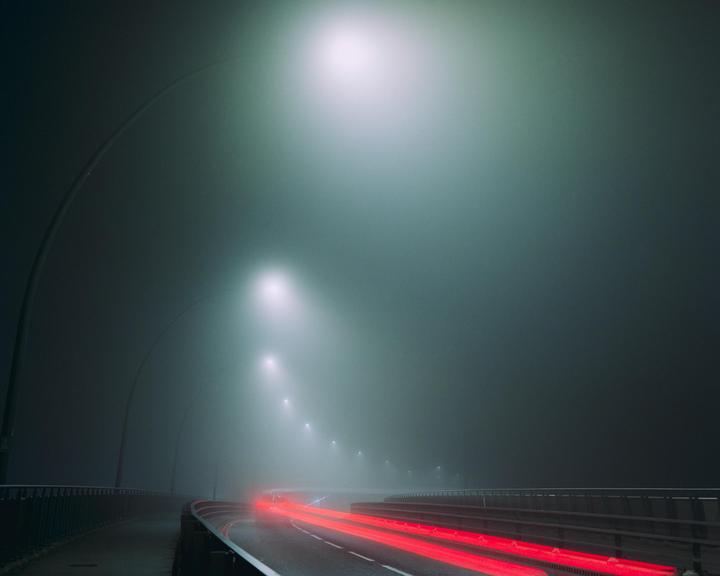 A foggy road at night is illuminated by streetlights and the red light trails of a vehicle.