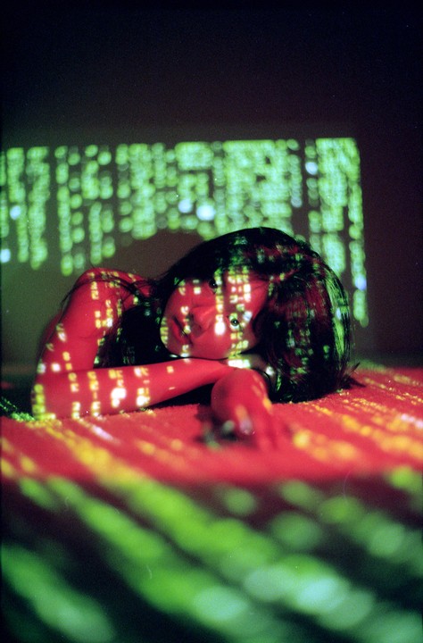 A woman lies on a red surface with green and red light patterns projected onto her and the surface.