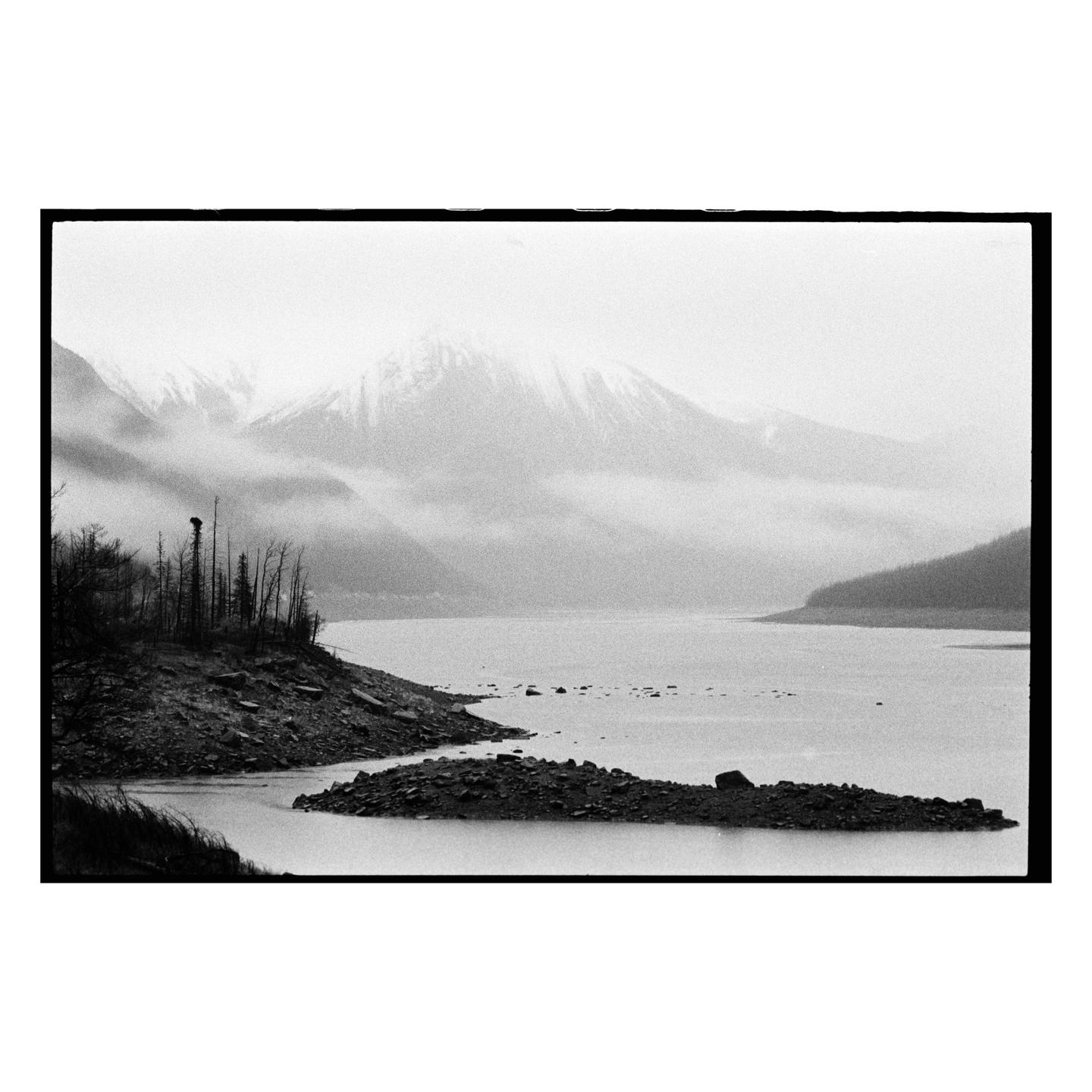 A body of water is surrounded by rocky shores and mountains covered in snow and fog.