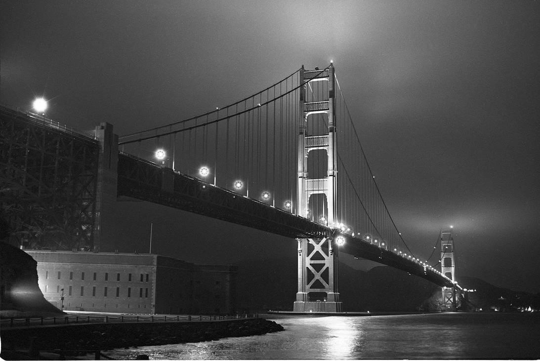 The Golden Gate Bridge is illuminated at night, with a building visible on the left shore and lights reflecting on the water.