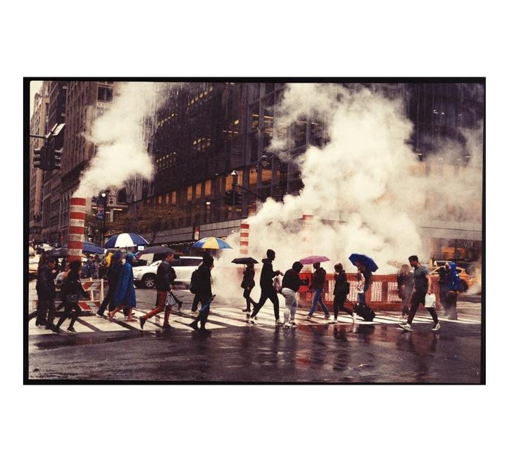 People with umbrellas cross a wet street with steam rising from manholes in a city.