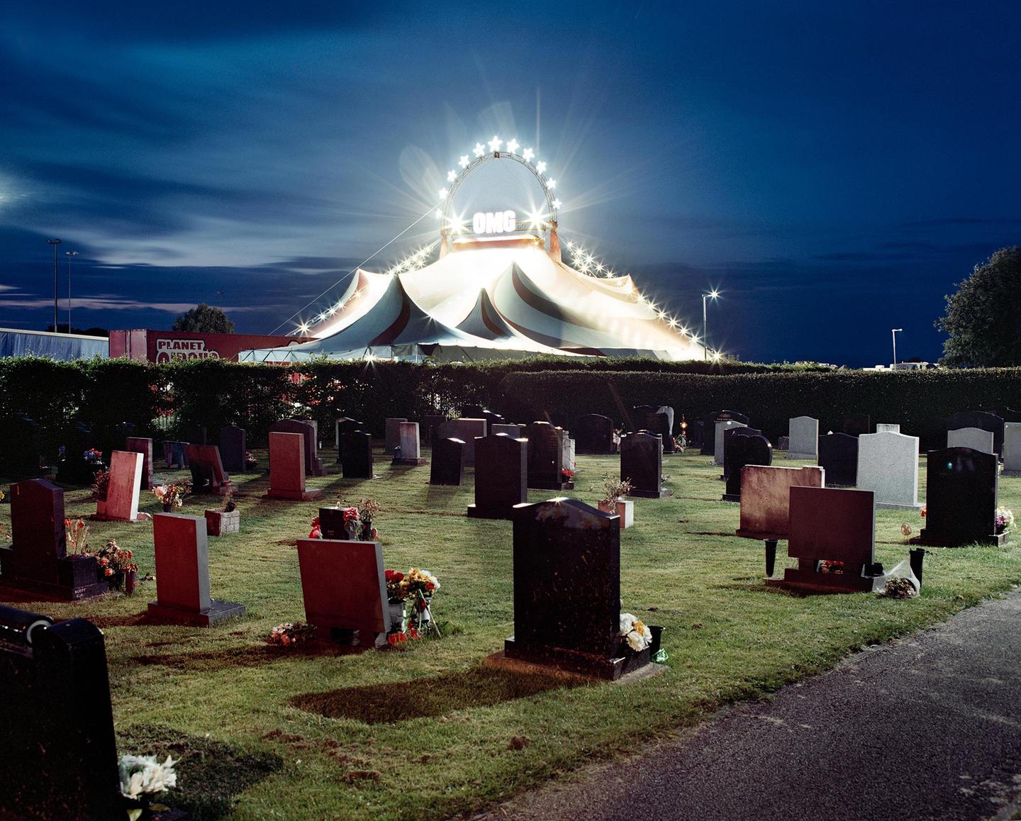 A brightly lit circus tent with a star-shaped sign reading "OMG" stands behind a hedge, overlooking a graveyard at night.