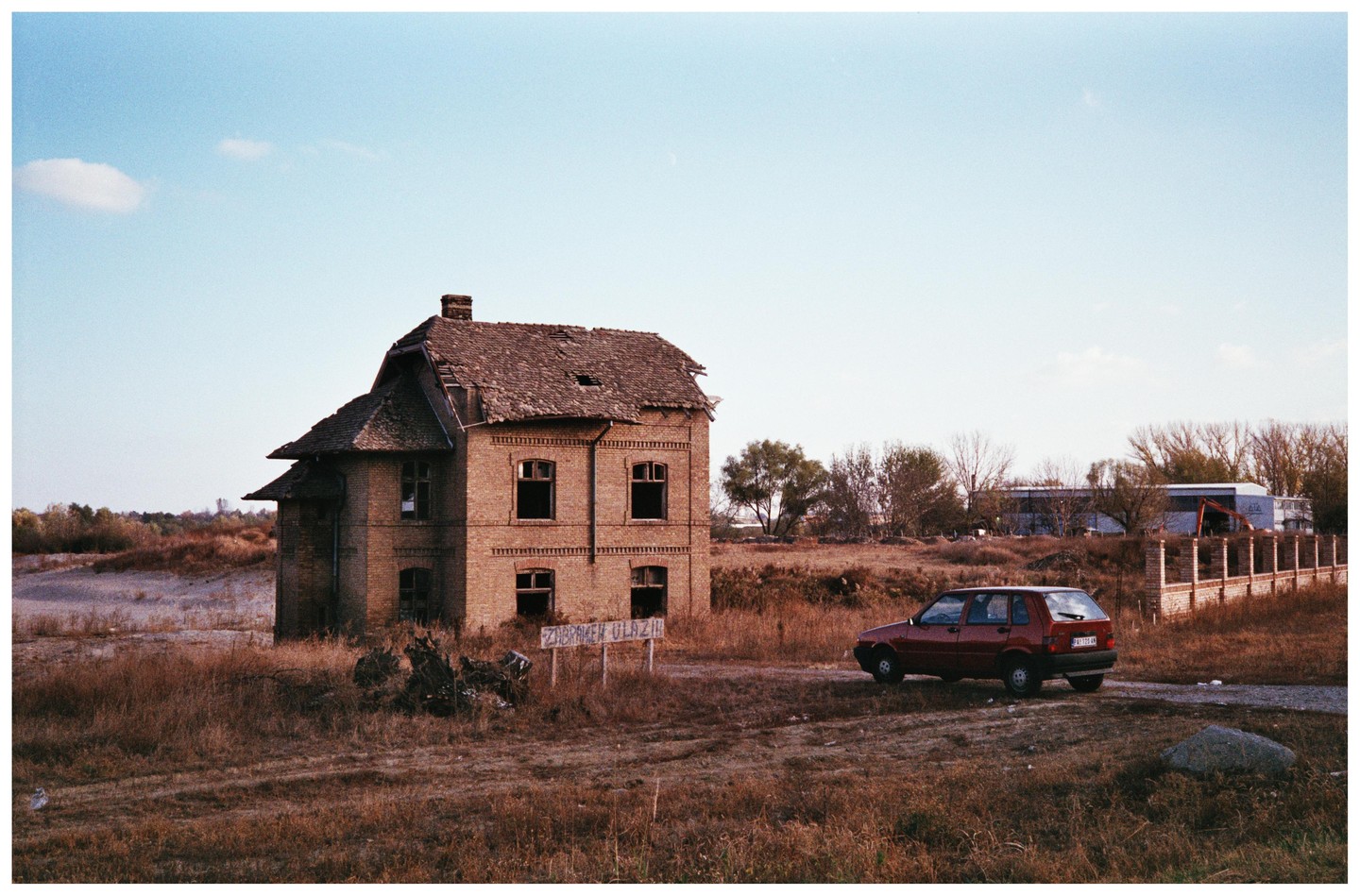 A red car is parked on a dirt road next to an abandoned brick building with a damaged roof.