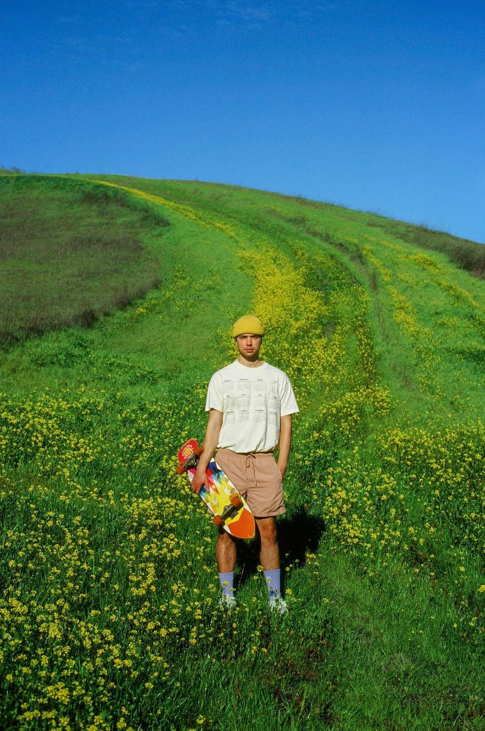 A man stands in a field of green grass and yellow flowers, holding a skateboard.