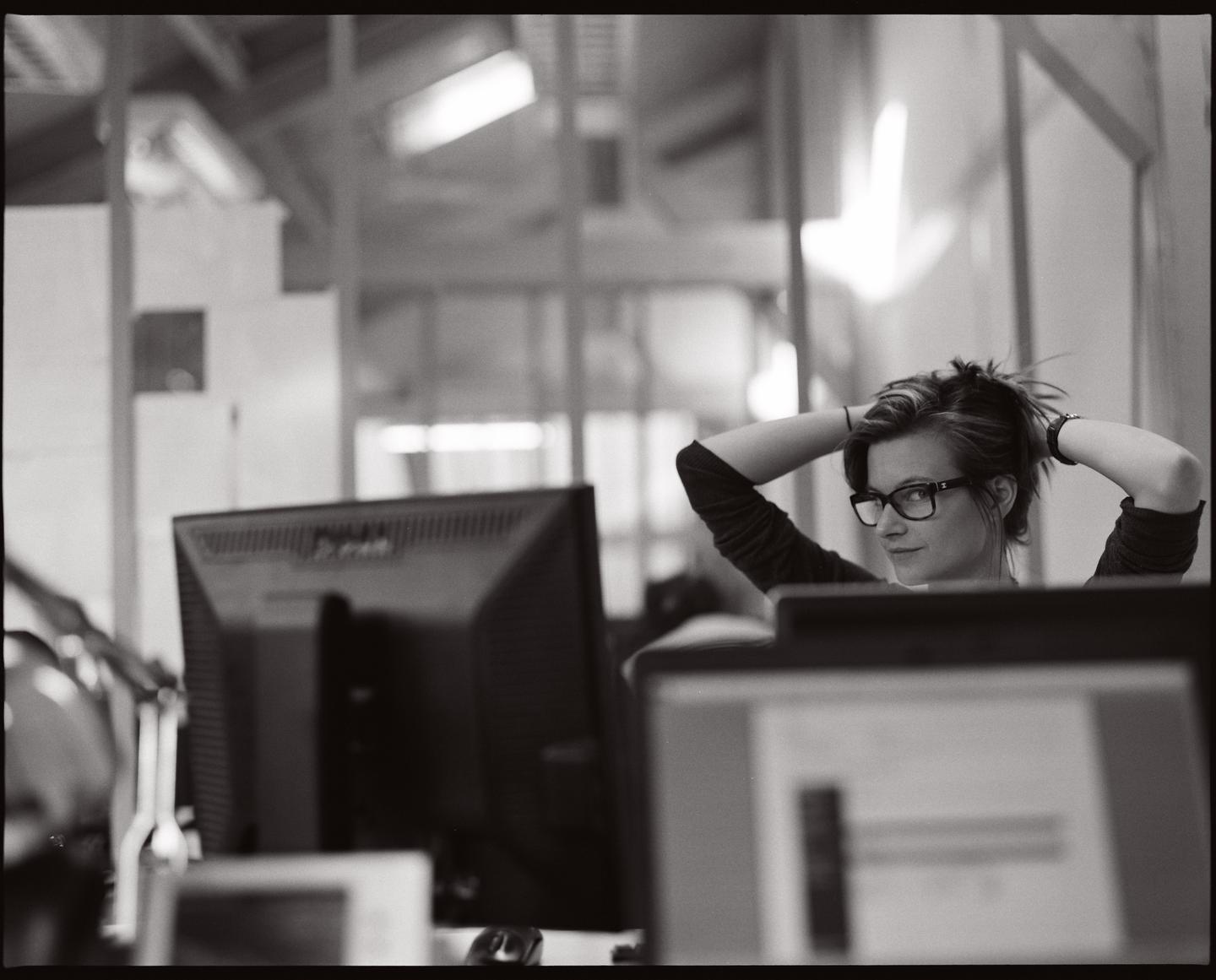A woman with glasses is sitting at a desk with her hands behind her head, looking over her shoulder at the viewer.