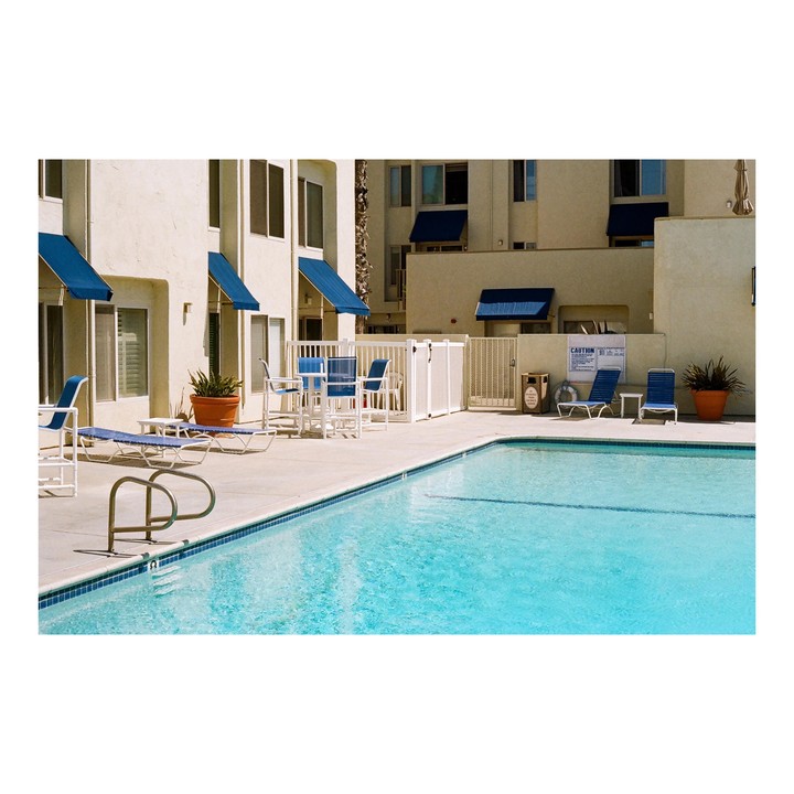 A swimming pool with blue lounge chairs and tables is next to a building with blue awnings.