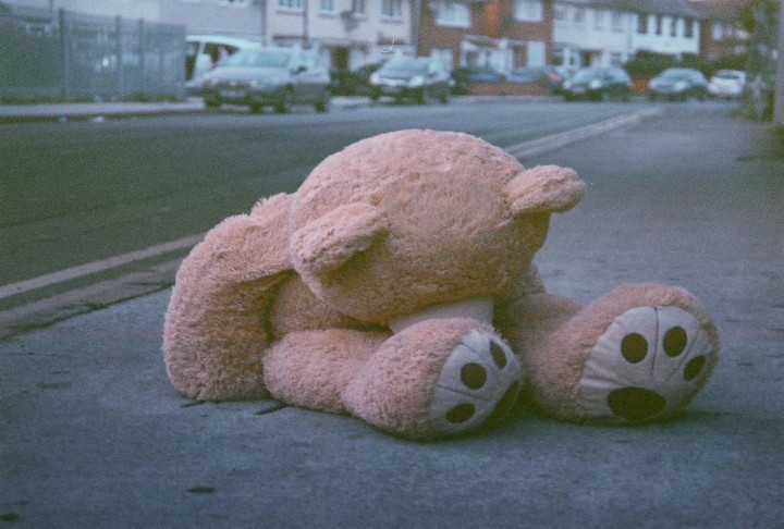 A large teddy bear sits on a sidewalk next to a street with cars and houses in the background.