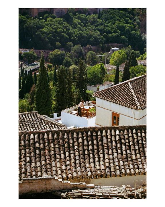 A man stands on a rooftop terrace overlooking a city with tiled roofs and lush green hills.