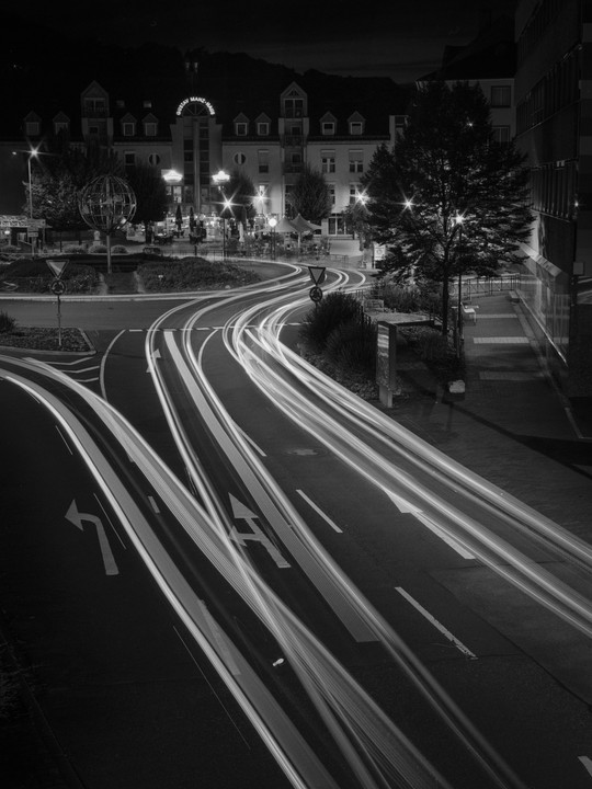 Light trails from vehicles illuminate a street at night, with buildings and trees visible in the background.