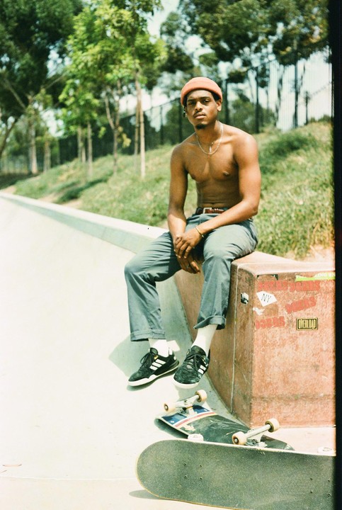 A man sits shirtless on a concrete ledge next to a skateboard at a skate park.