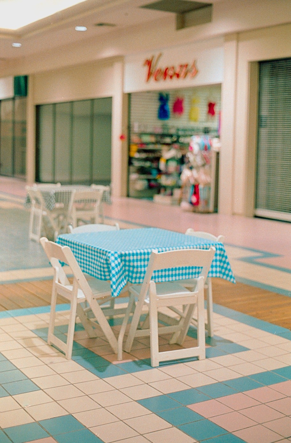 A blue and white checkered tablecloth covers a table with four chairs in a mall with a store named "Versus" in the background.