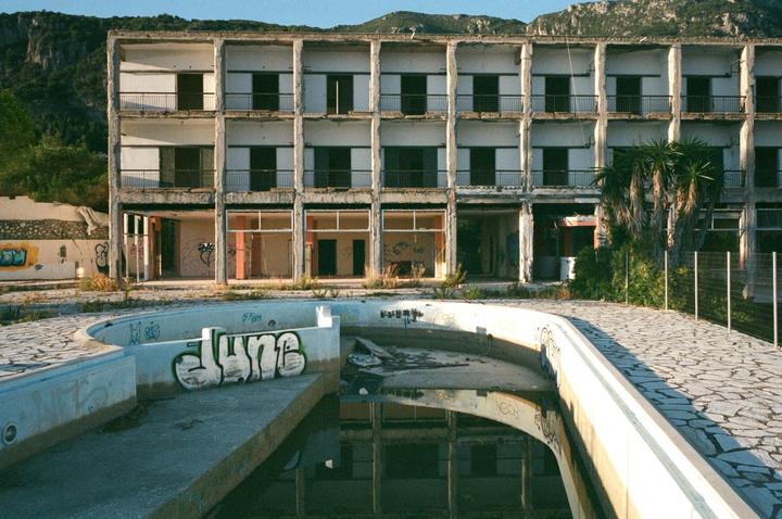 An abandoned hotel with a swimming pool in the foreground and mountains in the background is shown in a wide shot.
