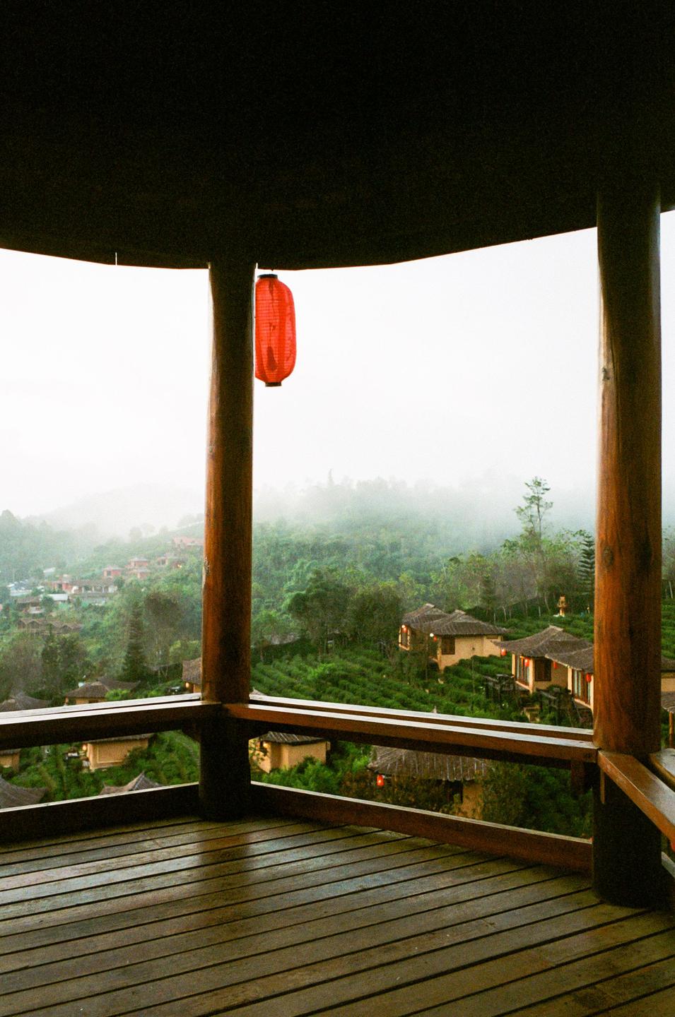 A red lantern hangs from a wooden structure overlooking a misty valley with houses and a tea farm.