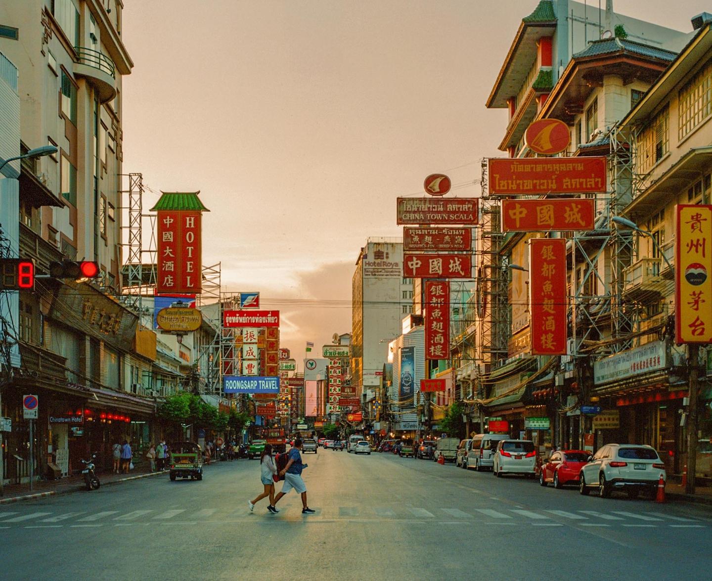 A couple crosses a busy street lined with buildings and signs in Bangkok.