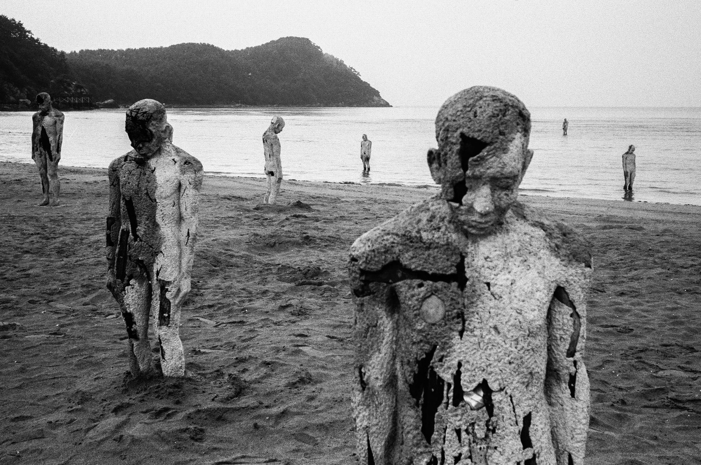 Several weathered human-shaped sculptures stand on a sandy beach with a calm sea and a tree-covered hill in the background.