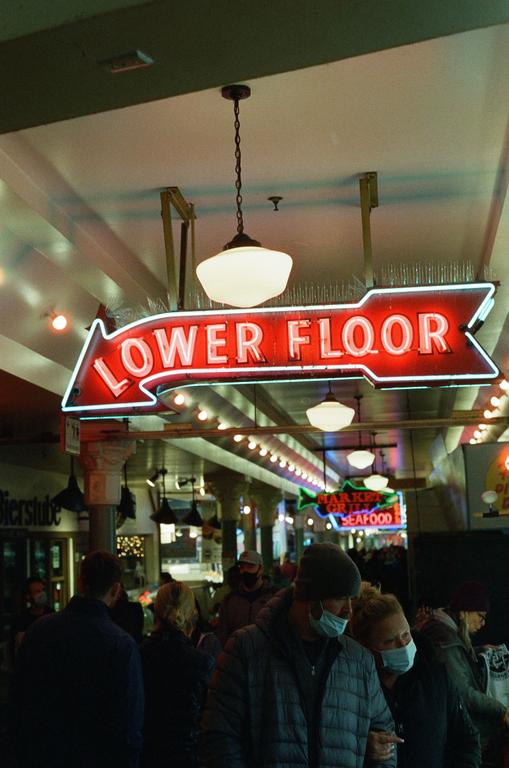 A red neon sign points to the lower floor of a market where people are walking.