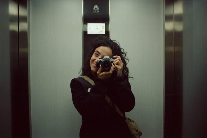 A woman with curly hair takes a self-portrait in an elevator, holding a camera to her face.