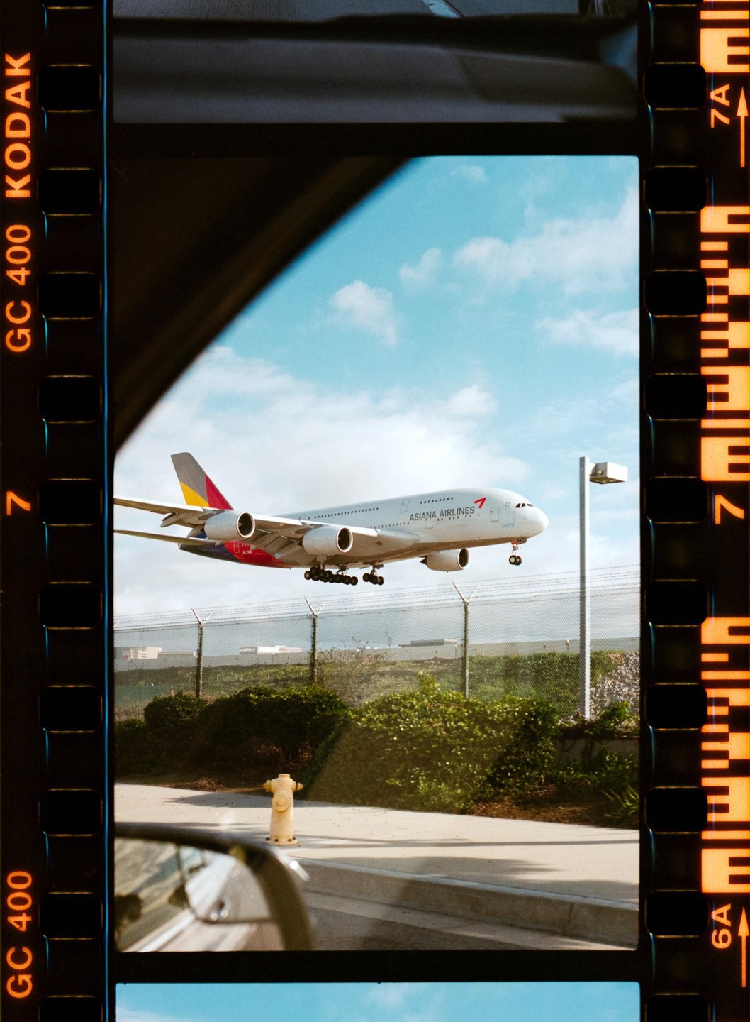 An Asiana Airlines airplane is captured mid-flight, seen through a car window with a fire hydrant in the foreground.