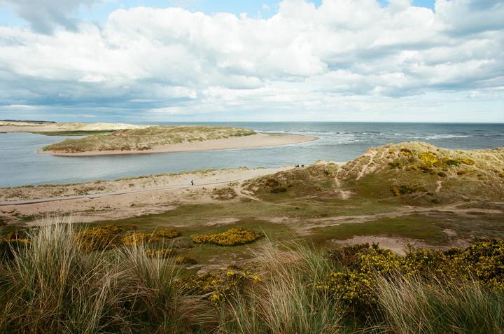 A wide view shows a river meeting the sea, with sand dunes and green vegetation under a cloudy sky.