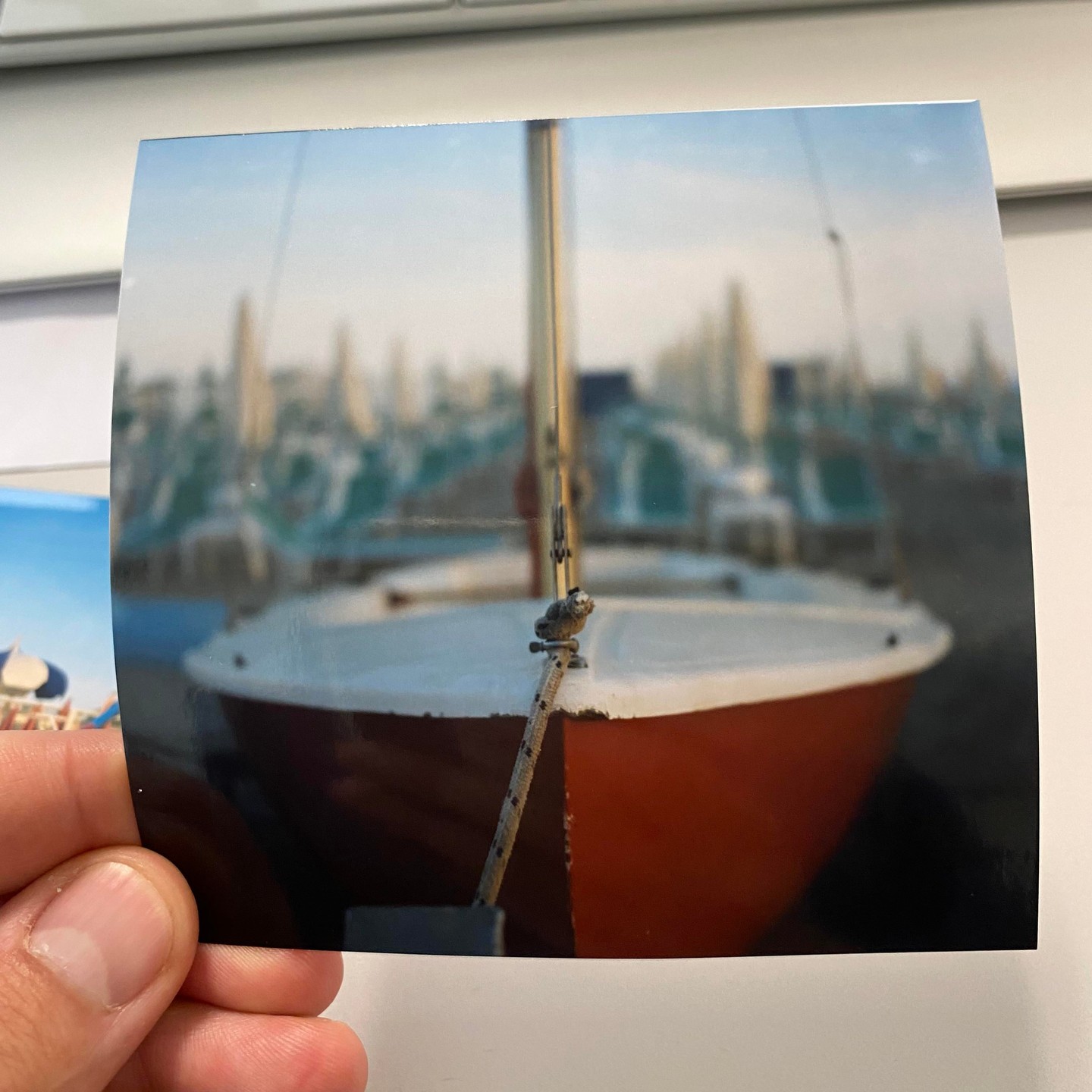 A small sailboat with a red hull and white deck is docked with other boats blurred in the background.