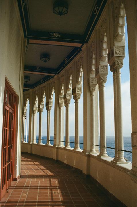 A long, arched balcony with ornate columns overlooks the ocean under a blue sky.