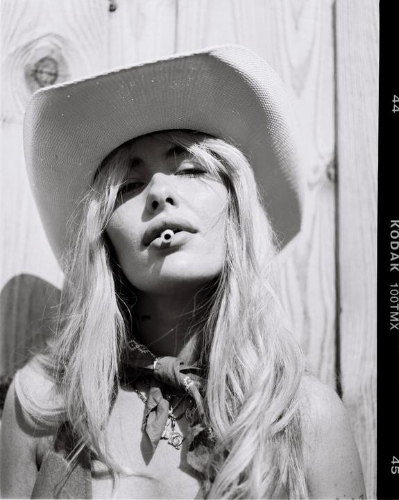 A woman wearing a cowboy hat and bandana holds a cigarette filter in her mouth.