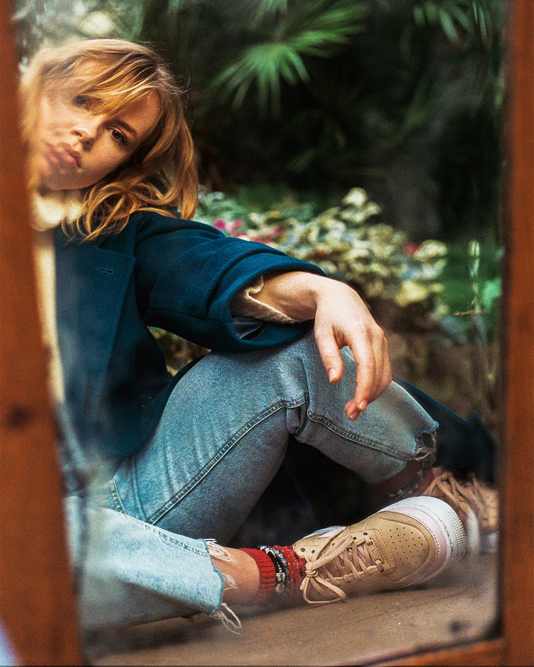 A woman with blonde hair and a blue coat sits on the ground, reflected in a mirror.