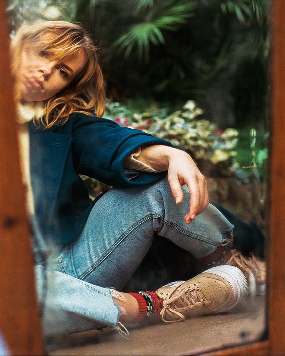 A woman with blonde hair and a blue coat sits on the ground, reflected in a mirror.