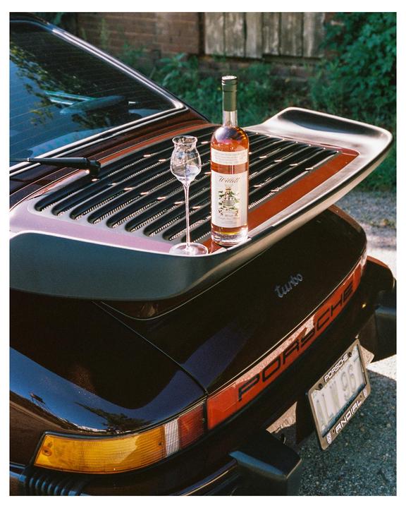 A bottle of liquor and a snifter glass sit on the spoiler of a black Porsche.