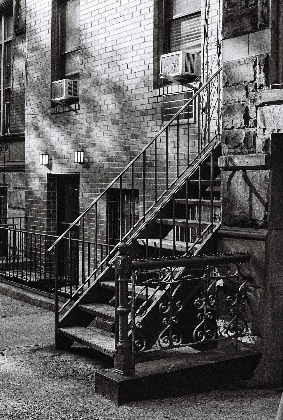 A black and white photo shows an exterior staircase with ornate railings leading up to a brick building with windows and air conditioning units.