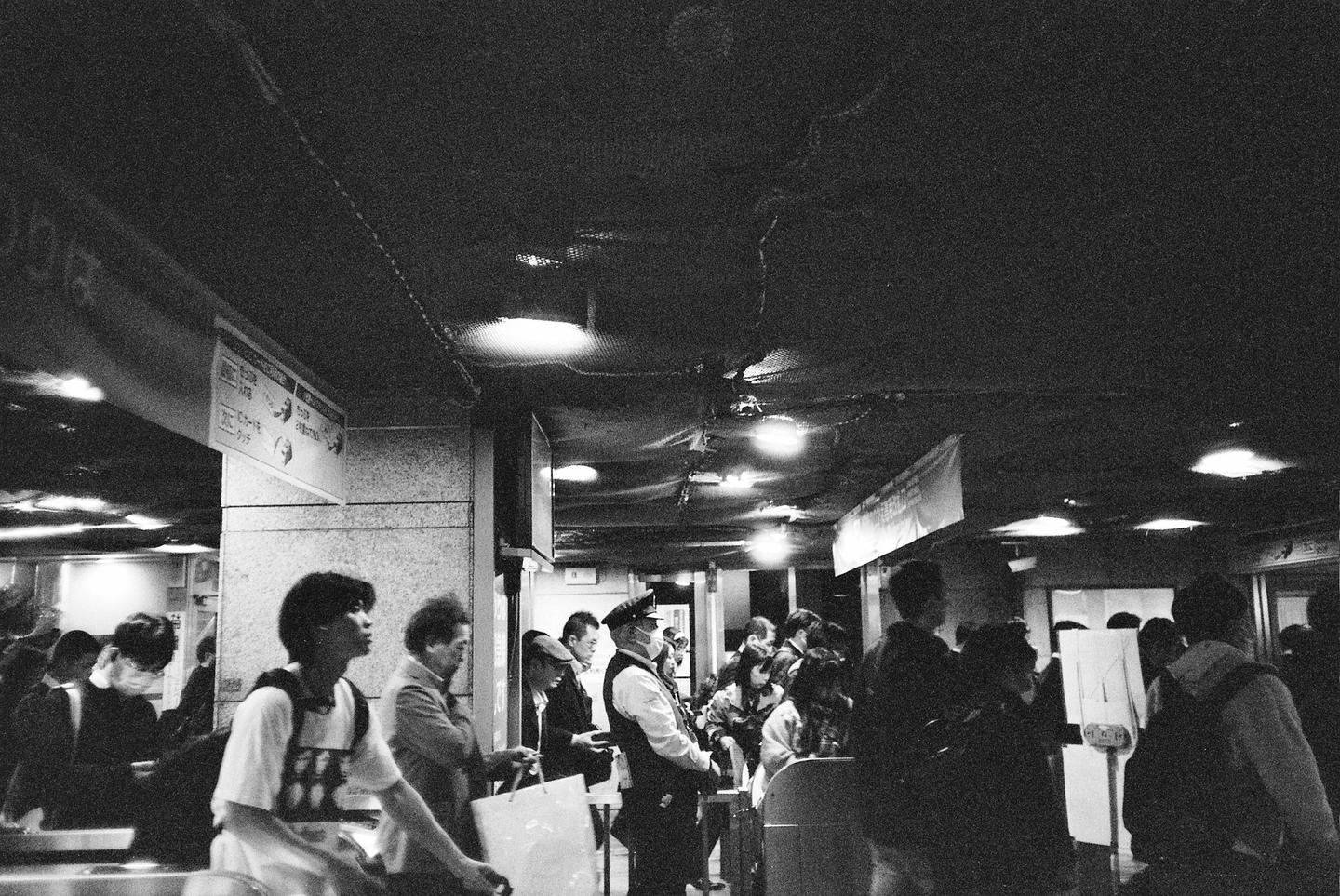 A crowd of people, including a man in a uniform, are gathered in a busy indoor area with overhead lights.