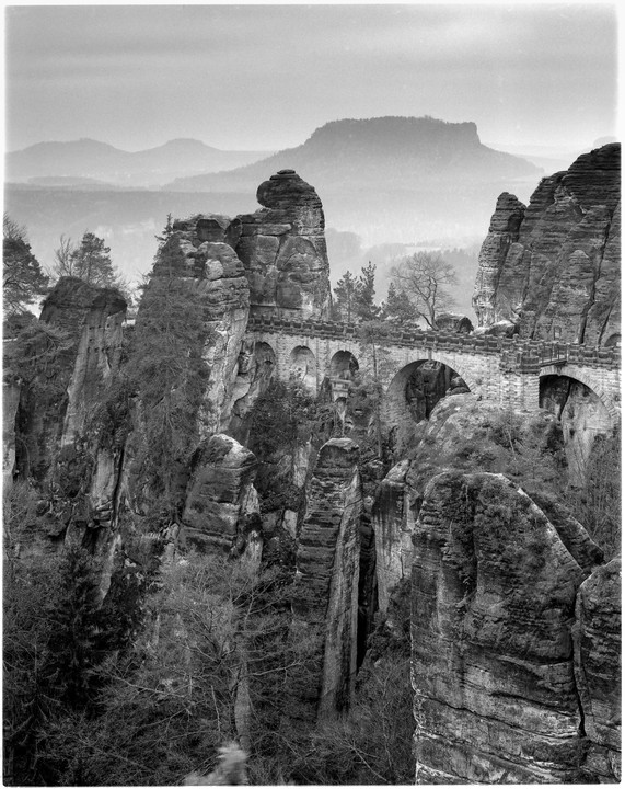 A stone bridge with multiple arches connects two large rock formations in a mountainous landscape.