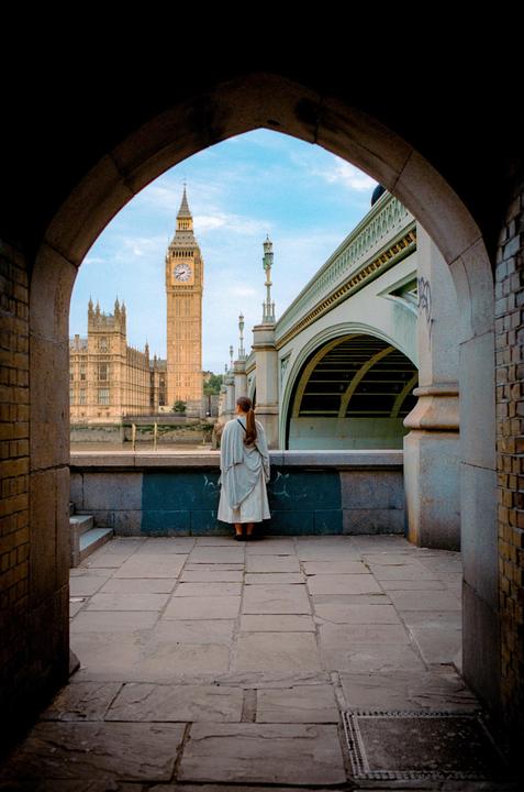 A woman stands with her back to the camera, looking at Big Ben and a bridge through an archway.