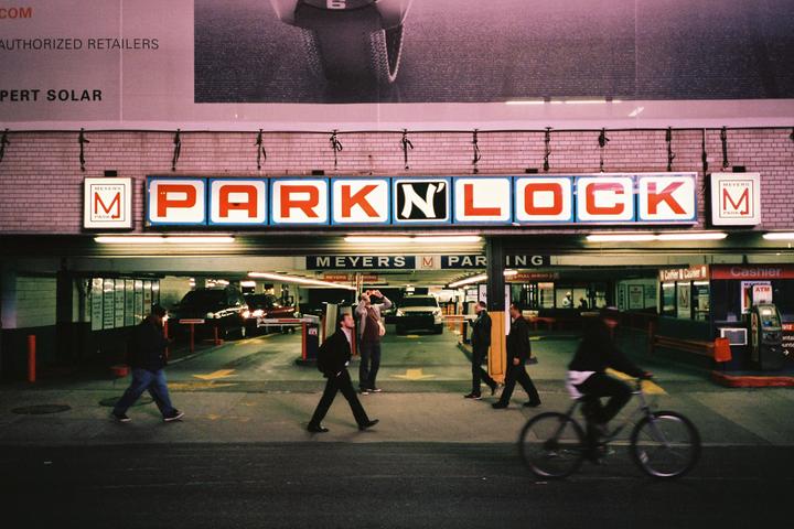 People walk and a person rides a bicycle past the entrance to a parking garage with a large "PARK N' LOCK" sign above it at night.