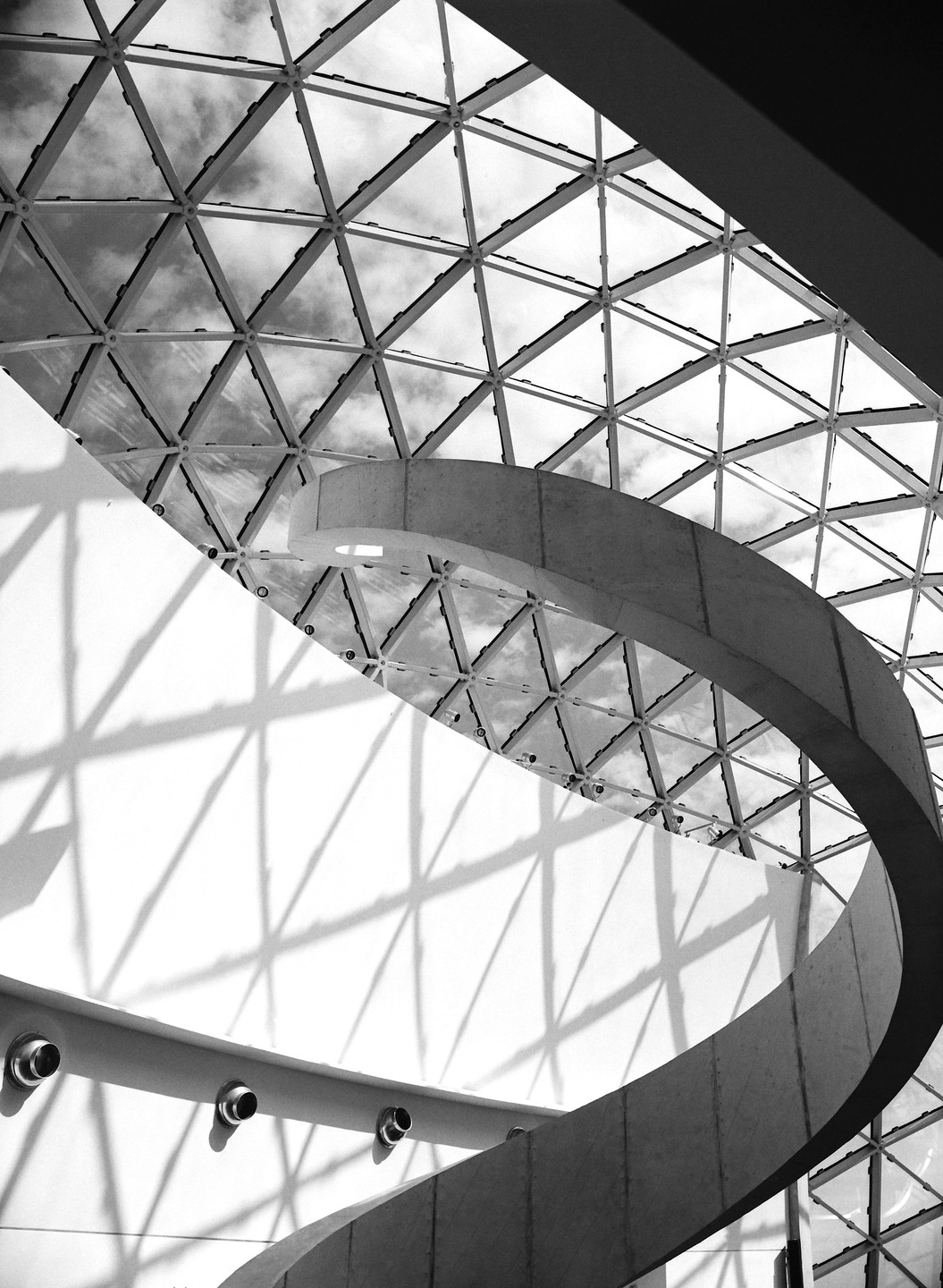 A spiral staircase curves upwards towards a geodesic dome ceiling, casting shadows on a white wall.