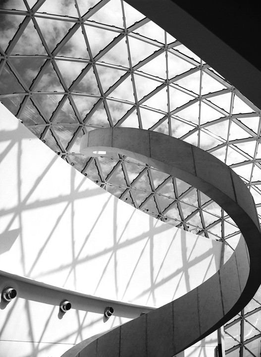 A spiral staircase curves upwards towards a geodesic dome ceiling, casting shadows on a white wall.