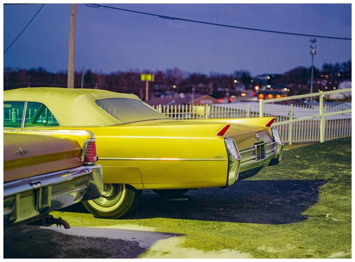 Two vintage cars are parked side by side at night, with a white fence and distant lights in the background.