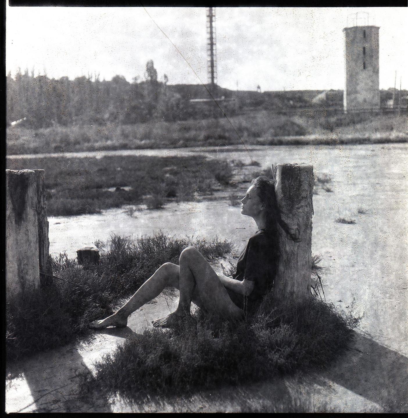 A woman sits barefoot in the grass, leaning against a wooden post, with a body of water and structures in the background.