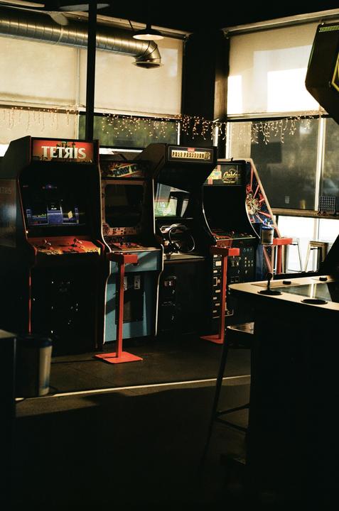 Several arcade game machines are lined up in a room with large windows and string lights.
