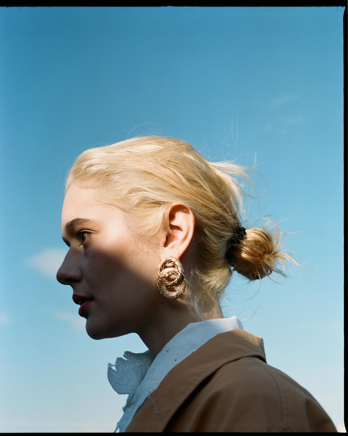 A woman with blonde hair in a bun and a large earring looks to the left against a clear blue sky.