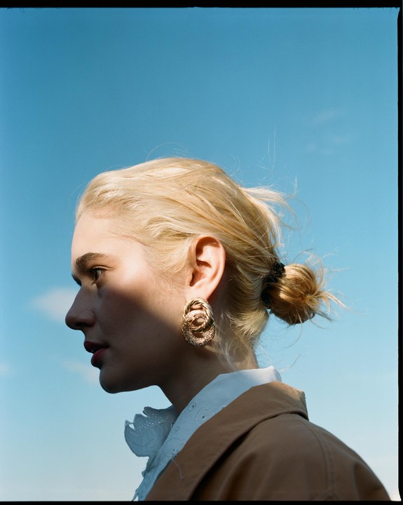 A woman with blonde hair in a bun and a large earring looks to the left against a clear blue sky.