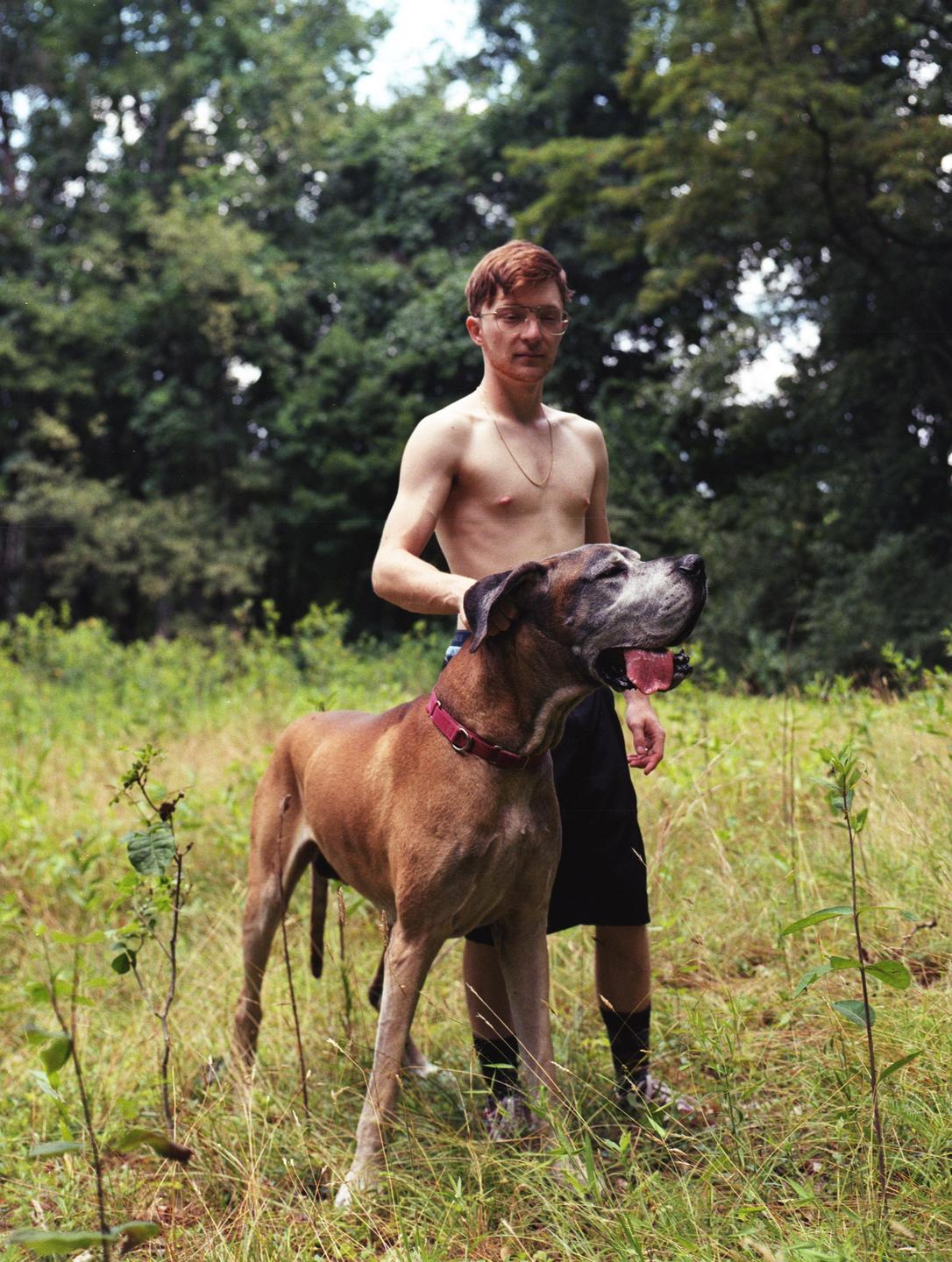 A man stands shirtless in a field with a large dog looking up at him.
