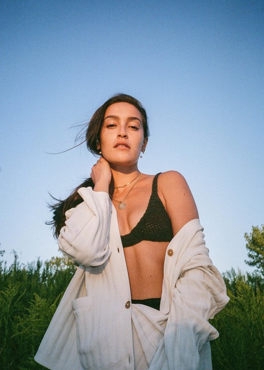 A woman wearing a white shirt and a black bra stands in a field under a clear sky.