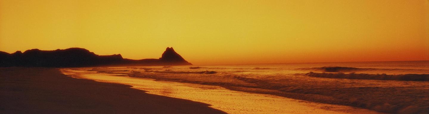 Waves crash on a beach with mountains in the background under an orange sky.
