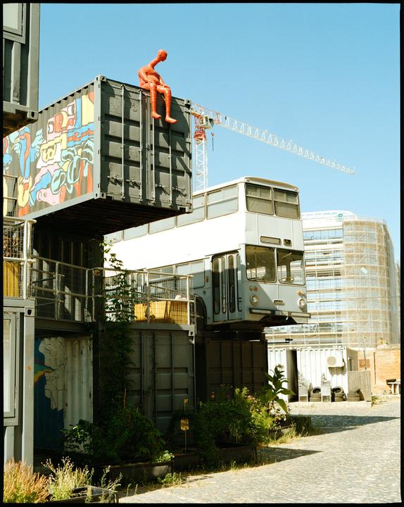 A red sculpture of a man sits atop a stack of shipping containers next to a bus.