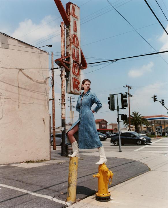 A woman in a denim dress and white boots stands on a yellow pole and fire hydrant next to a liquor sign.