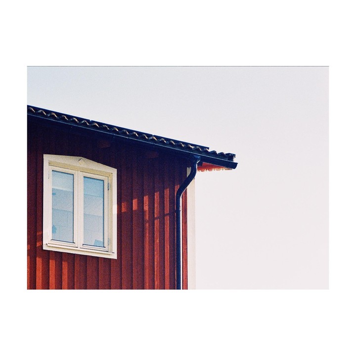 The side of a red house with a white window and a dark roof is shown against a bright sky.