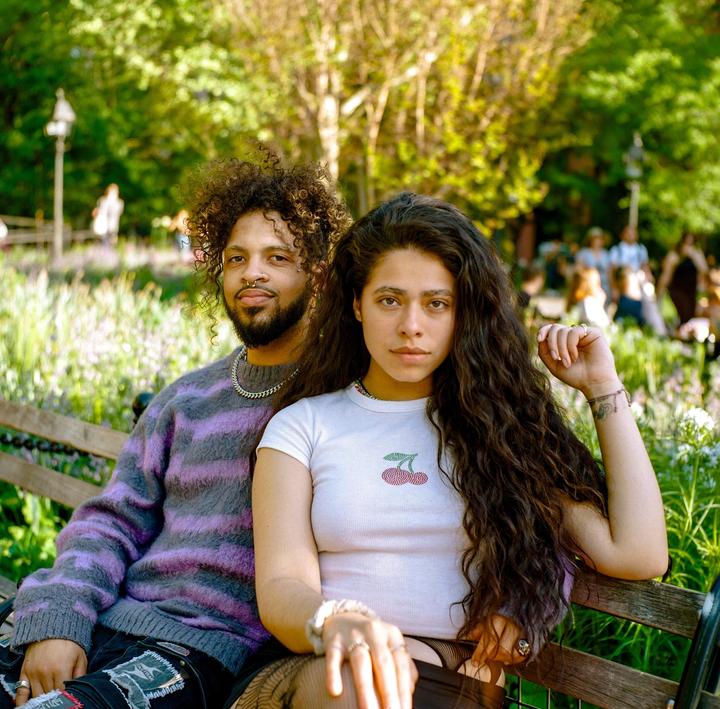 A couple sits on a park bench surrounded by green foliage and other people in the background.