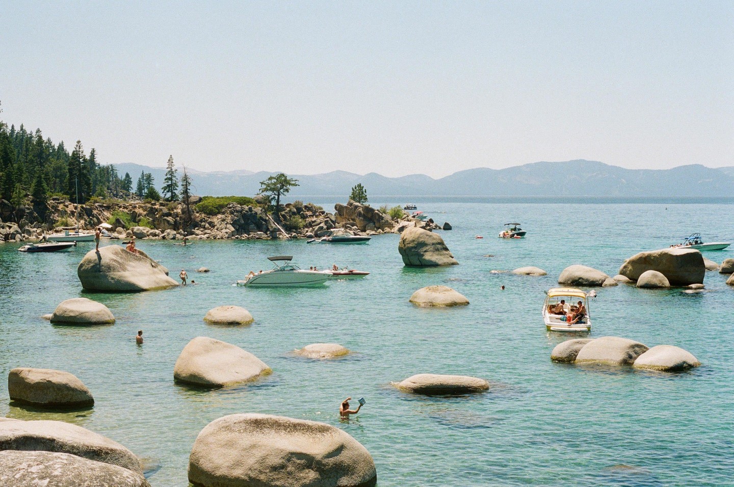 Boats and people are in a clear blue lake surrounded by large rocks and a forested shore.
